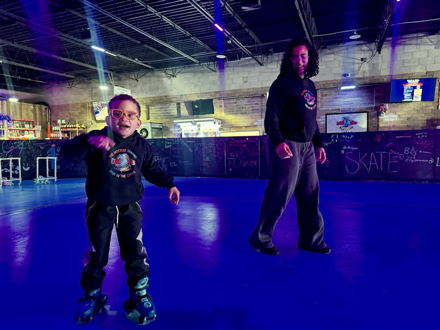 A young skater in orange glasses skating with a coach behind him